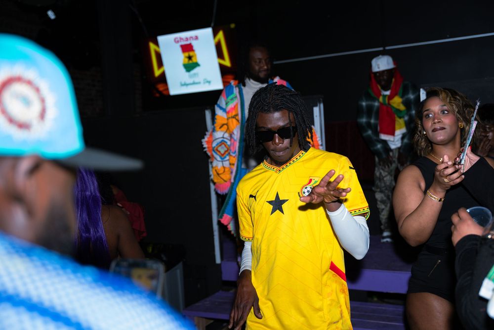 Man in yellow Ghana soccer jersey and sunglasses gestures at a lively indoor event with colorful decorations.