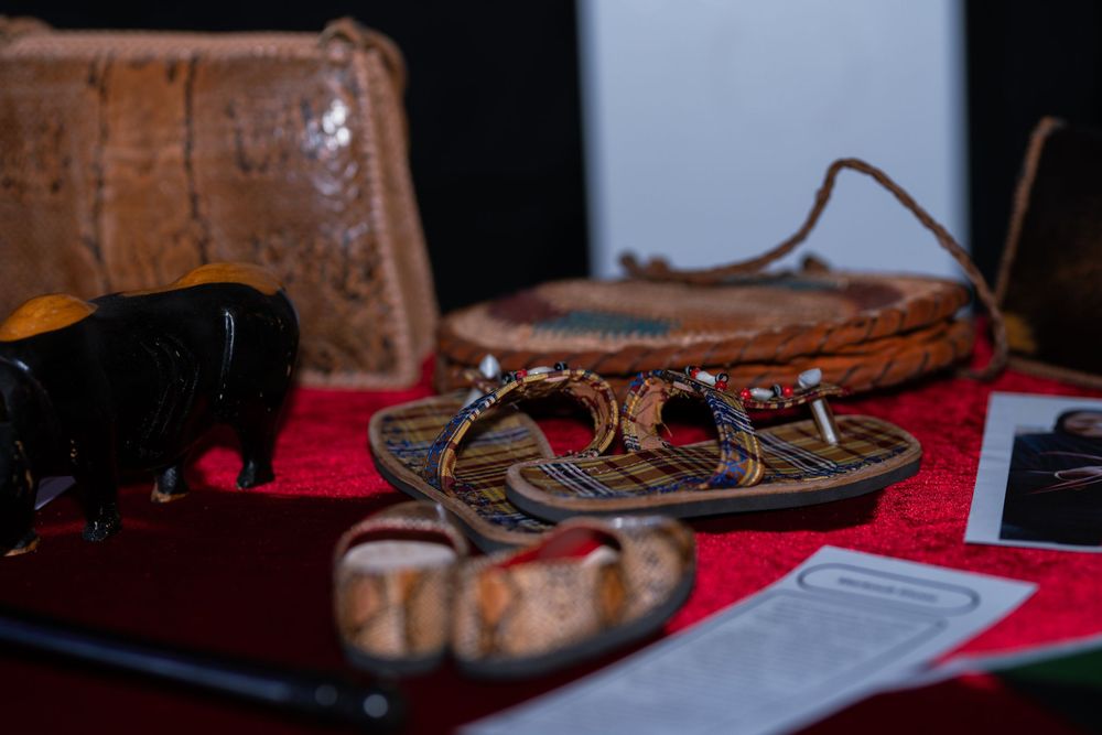 Handcrafted leather sandals, bag, and tribal figurines displayed on red cloth at a cultural market.