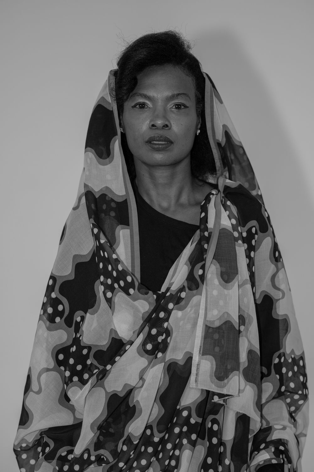 Black and white portrait of a woman draped in a patterned fabric wrap over her head and shoulders.