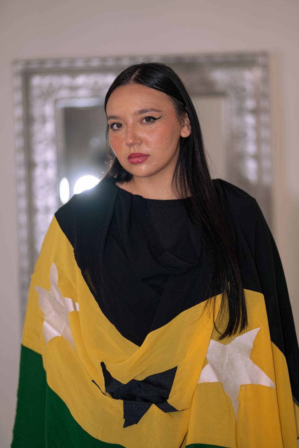 Young woman draped in a green, black, and white starred flag poses indoors near a mirror.