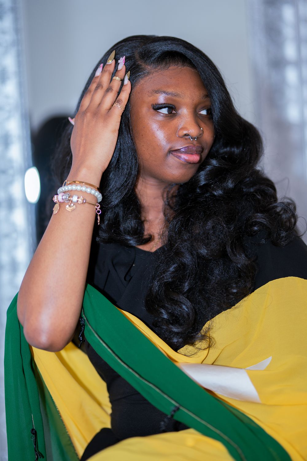 Young Black woman with long wavy hair holds head gracefully, wearing bracelets and black top with green and yellow fabric.
