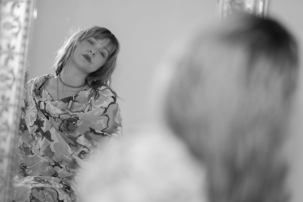 Black and white photo of a blonde woman in a floral dress gazing at her reflection in an ornate mirror.