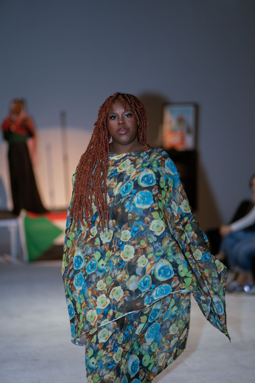 Model walks runway in flowing blue floral print kaftan dress with long red braids.