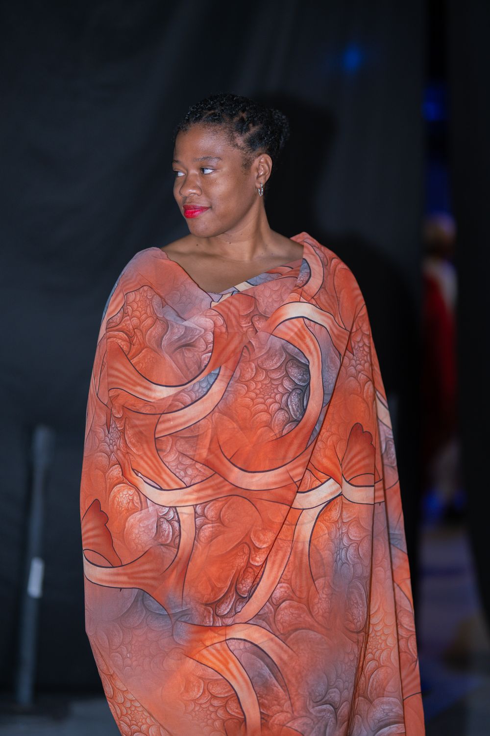 Black woman models stunning orange patterned wrap garment with intricate swirl design at a fashion showcase event.