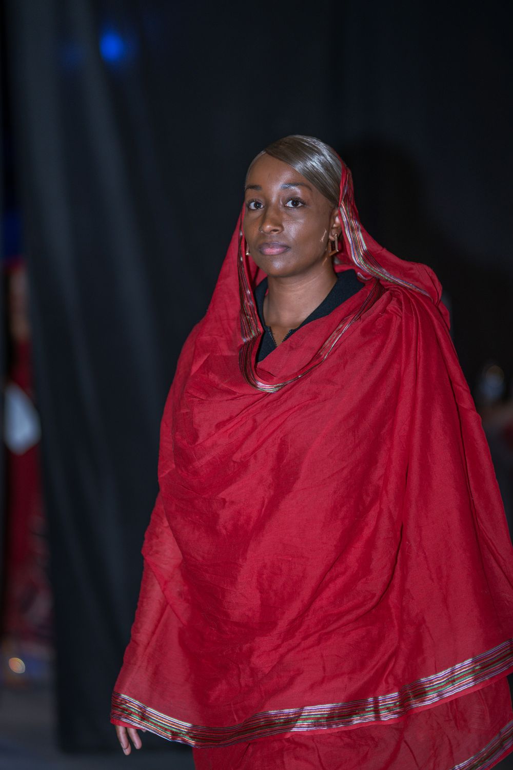 Woman in elegant red traditional draped fabric with gold trim walks confidently on a dark stage.