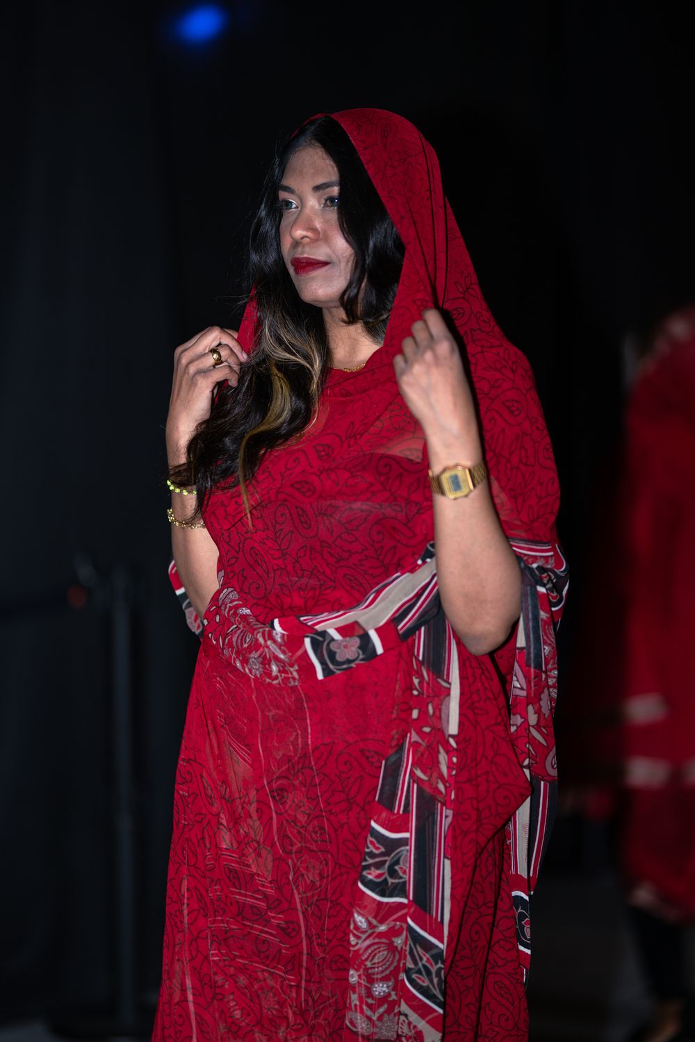Woman in elegant red lace hooded dress and gold jewelry posing against a dark background.