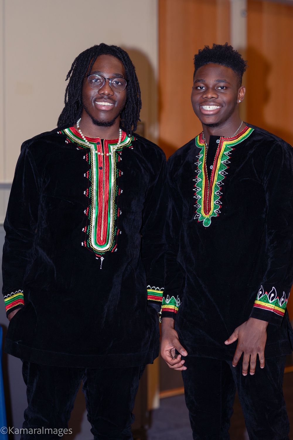 Two young men smiling in matching black velvet African traditional attire with colorful red, green, and gold embroidery.
