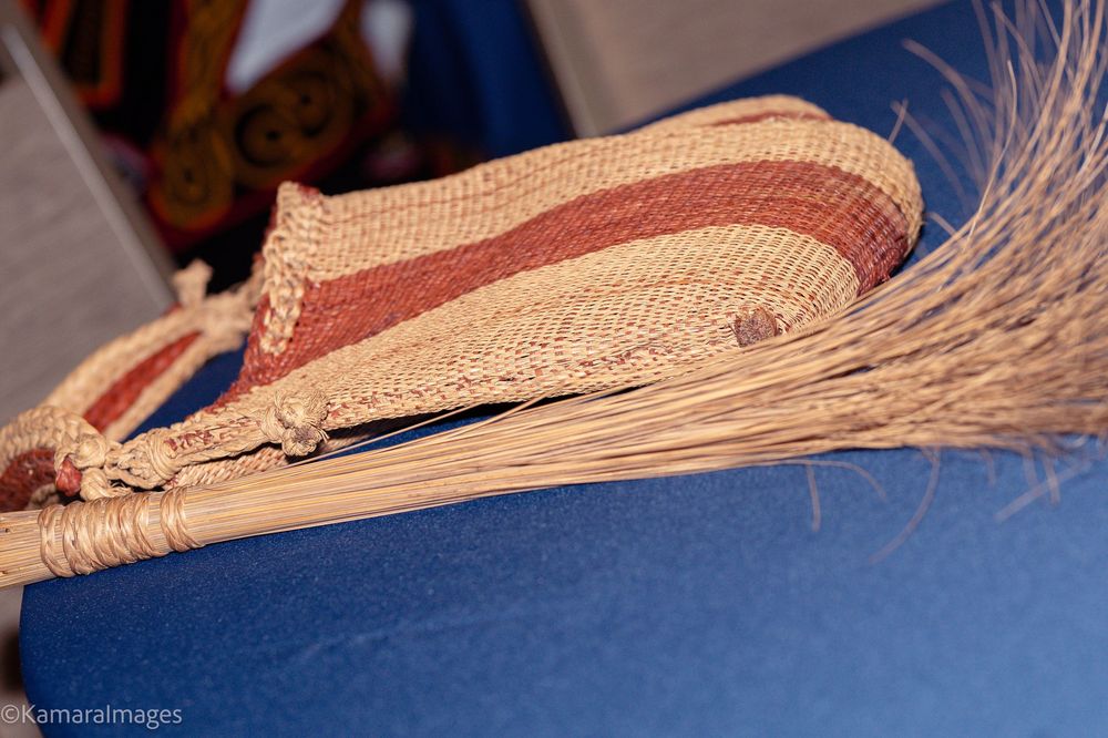 Handwoven straw basket with brown stripe pattern resting on dried grass broom on blue fabric surface.