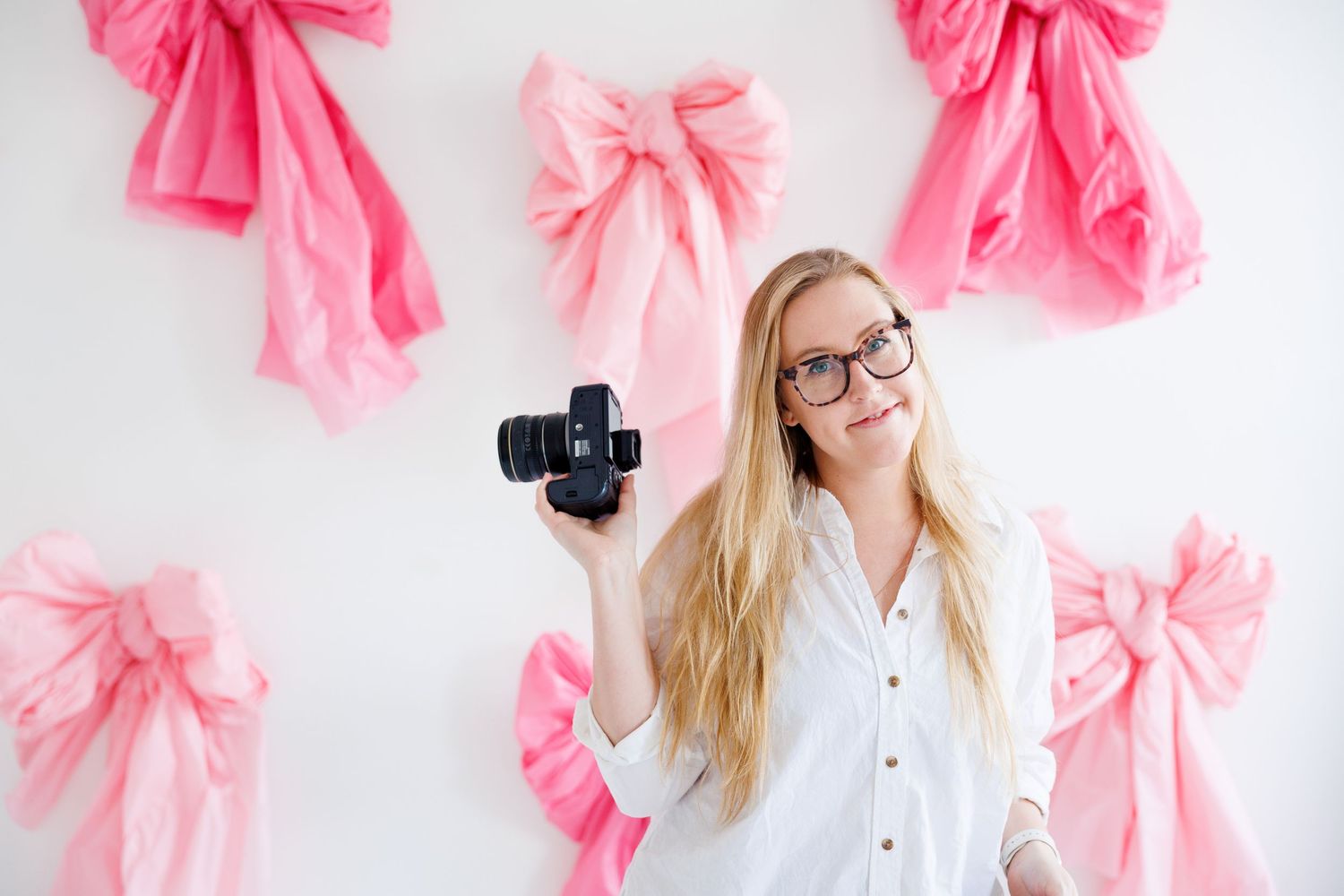 A person in a white shirt smiles while working with a camera against a pink bow decorated backdrop.