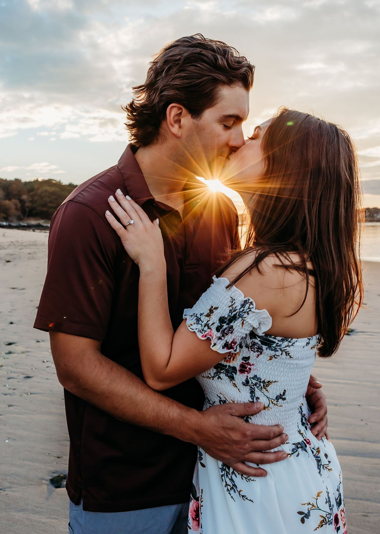 A couple embraces in casual attire against a sunlit outdoor backdrop with wooden fencing and greenery.