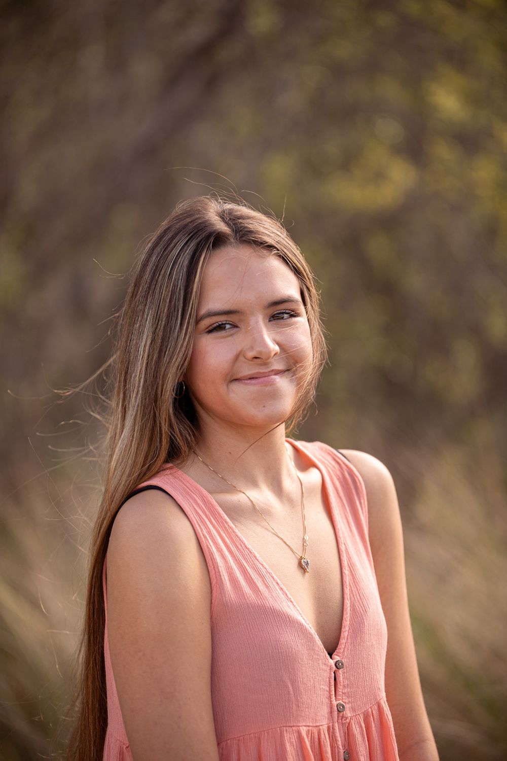 A person in a coral sleeveless dress poses outdoors with natural lighting and bokeh background.