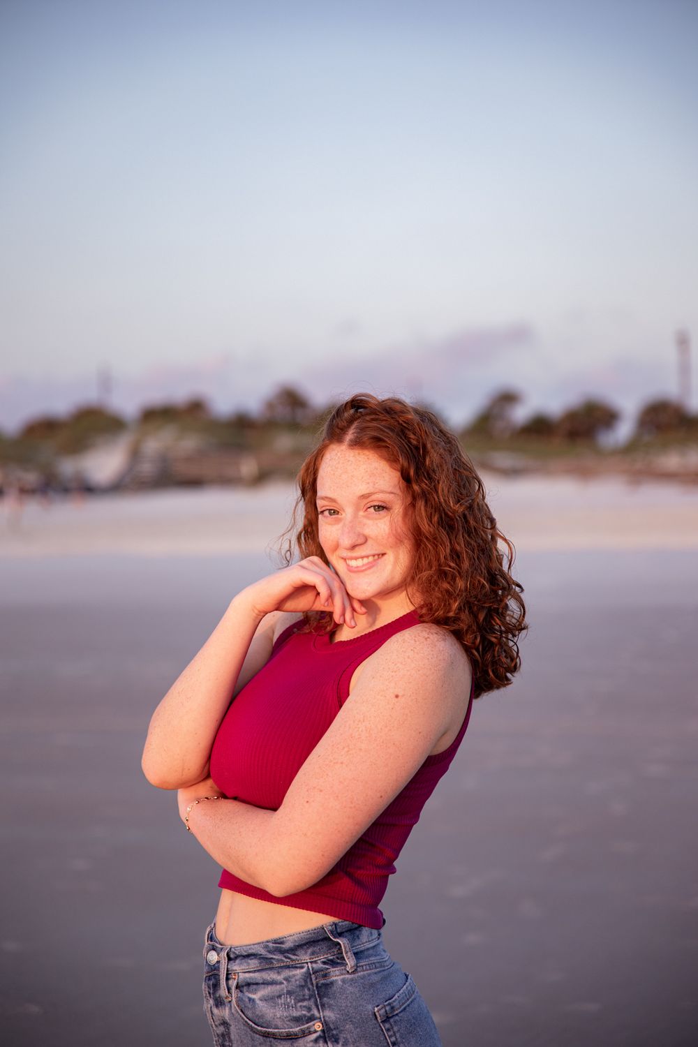 A person in a red top and jeans poses playfully on a beach at sunset.