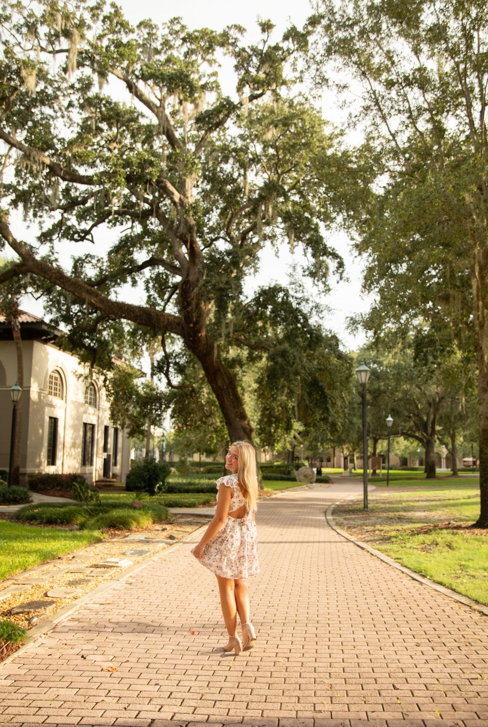 A person in a floral dress stands on a brick pathway surrounded by large oak trees and Spanish architecture.