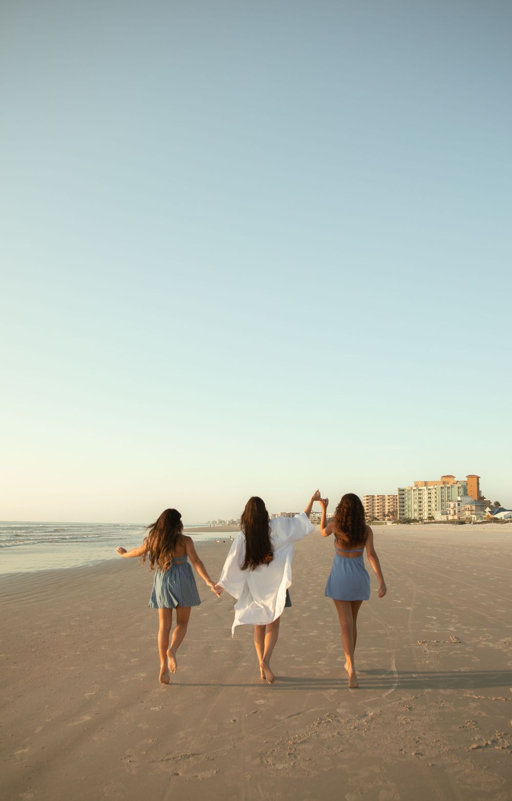 Three friends walking hand in hand on a beach at sunset with condos in the background.