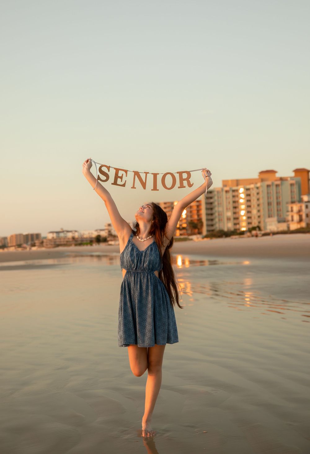 Person in blue dress twirling joyfully on beach at sunset with coastal buildings in background.