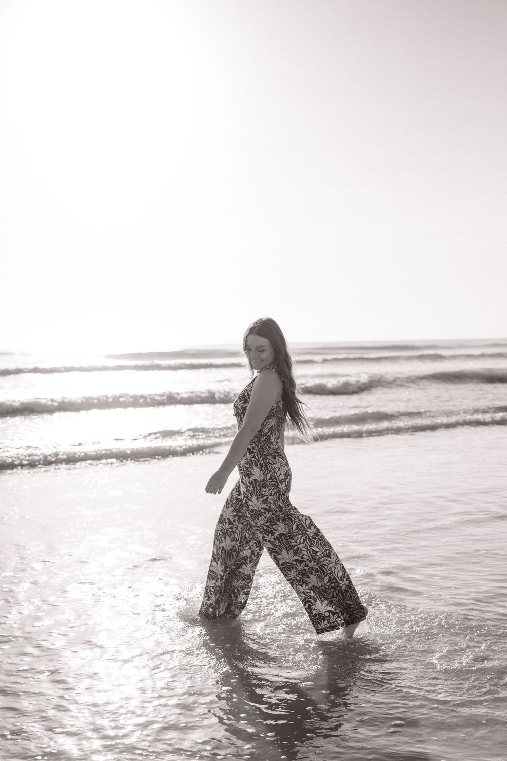 Person in patterned jumpsuit walking through ocean waves in black and white.