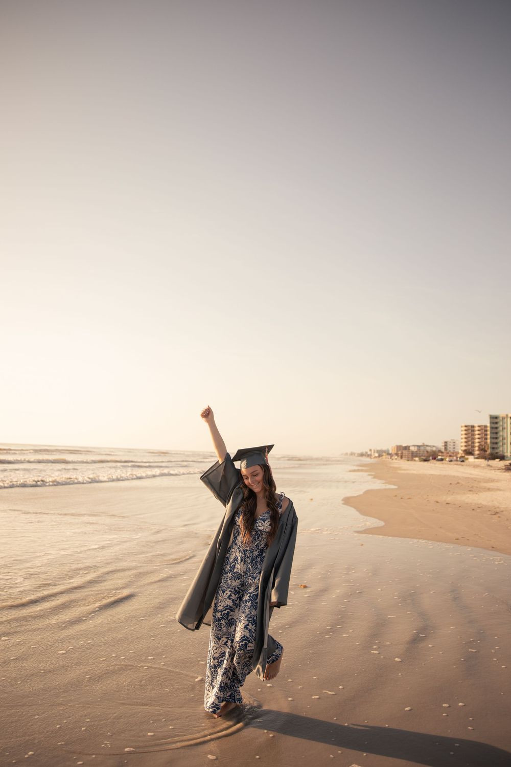 Person in graduation gown celebrating on beach at sunset.