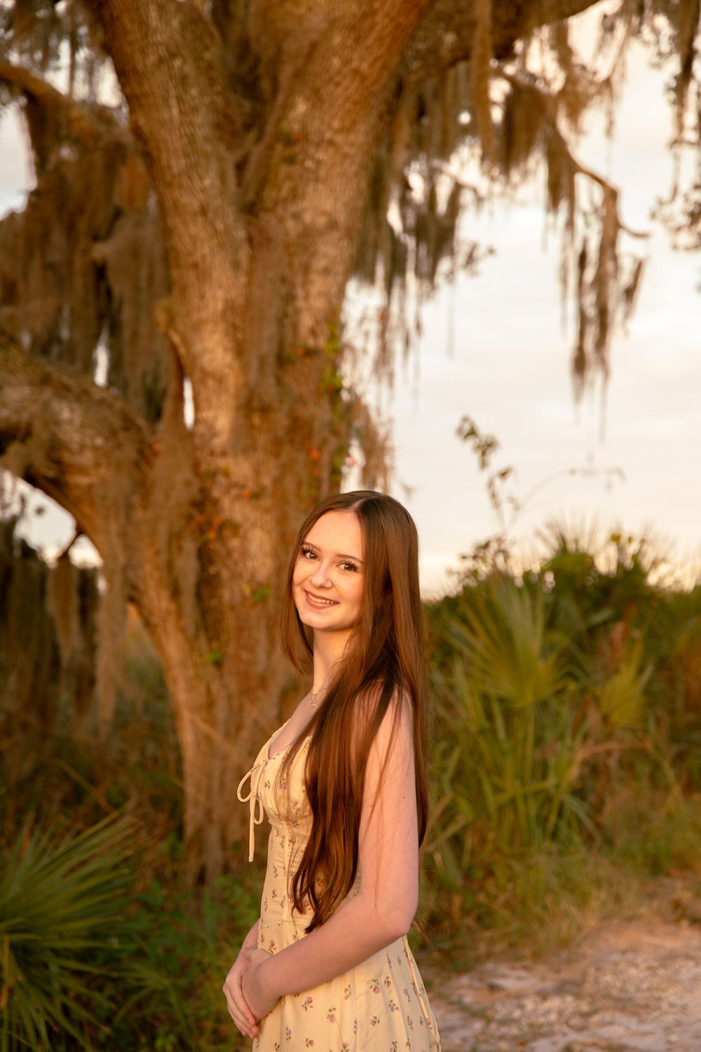 A person in a pink dress poses outdoors against Spanish moss-draped trees during golden hour sunset lighting.