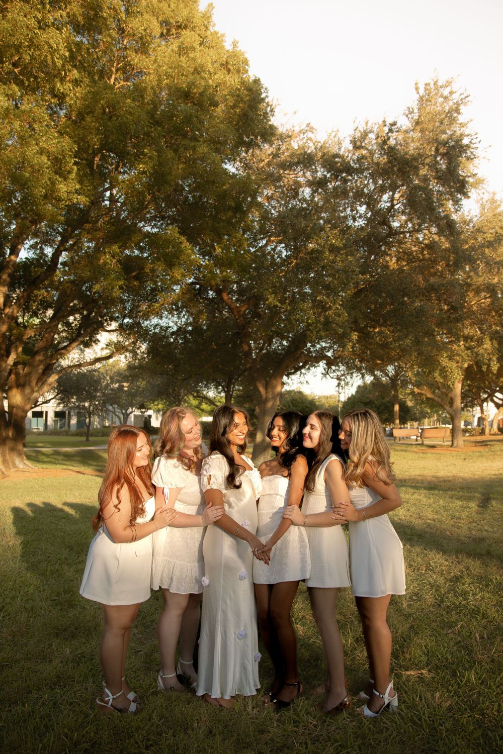 A group of graduates in white dresses poses together in a park during golden hour.