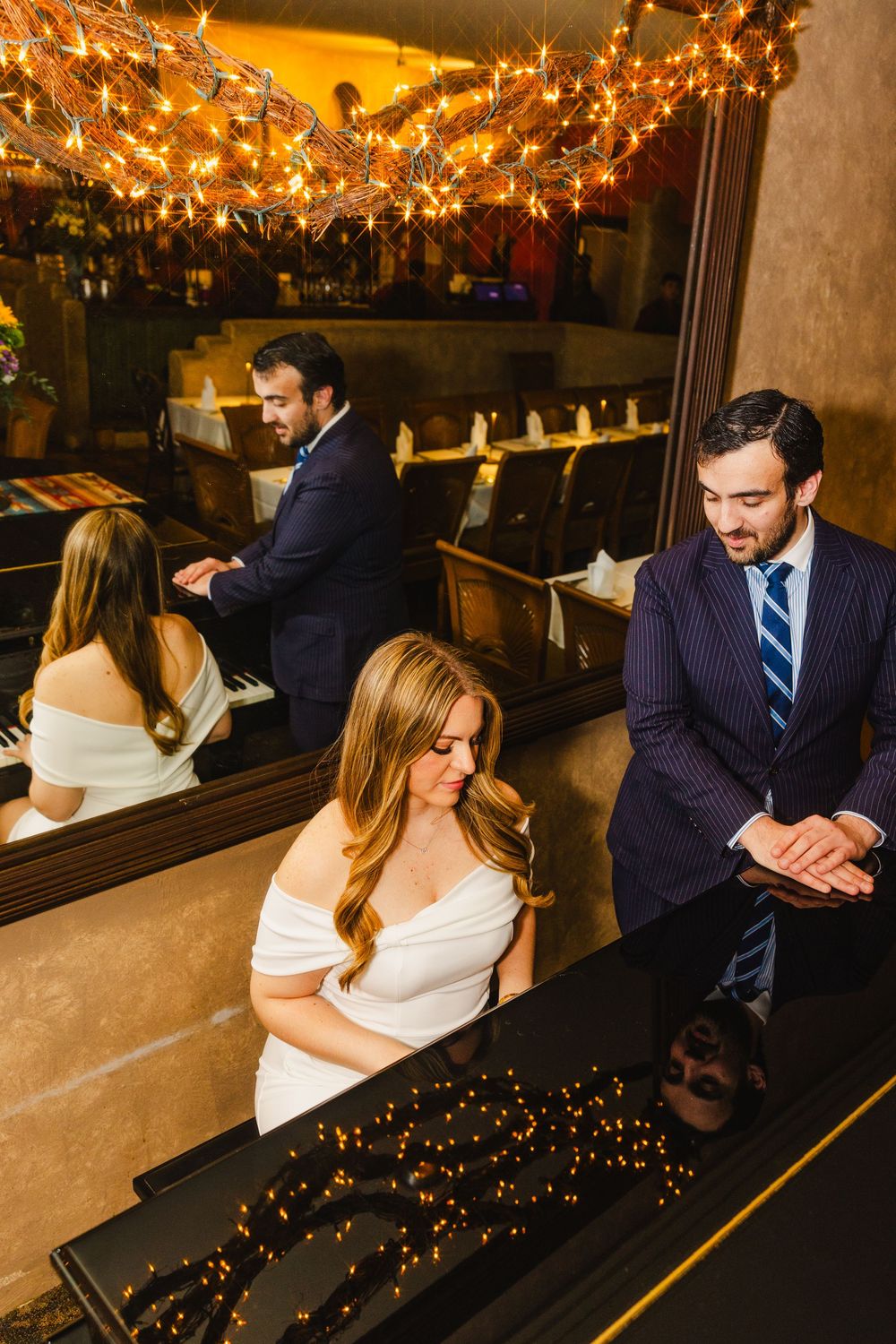 A group of well-dressed people enjoying drinks and conversation in a dimly lit bar or restaurant with string lights overhead.