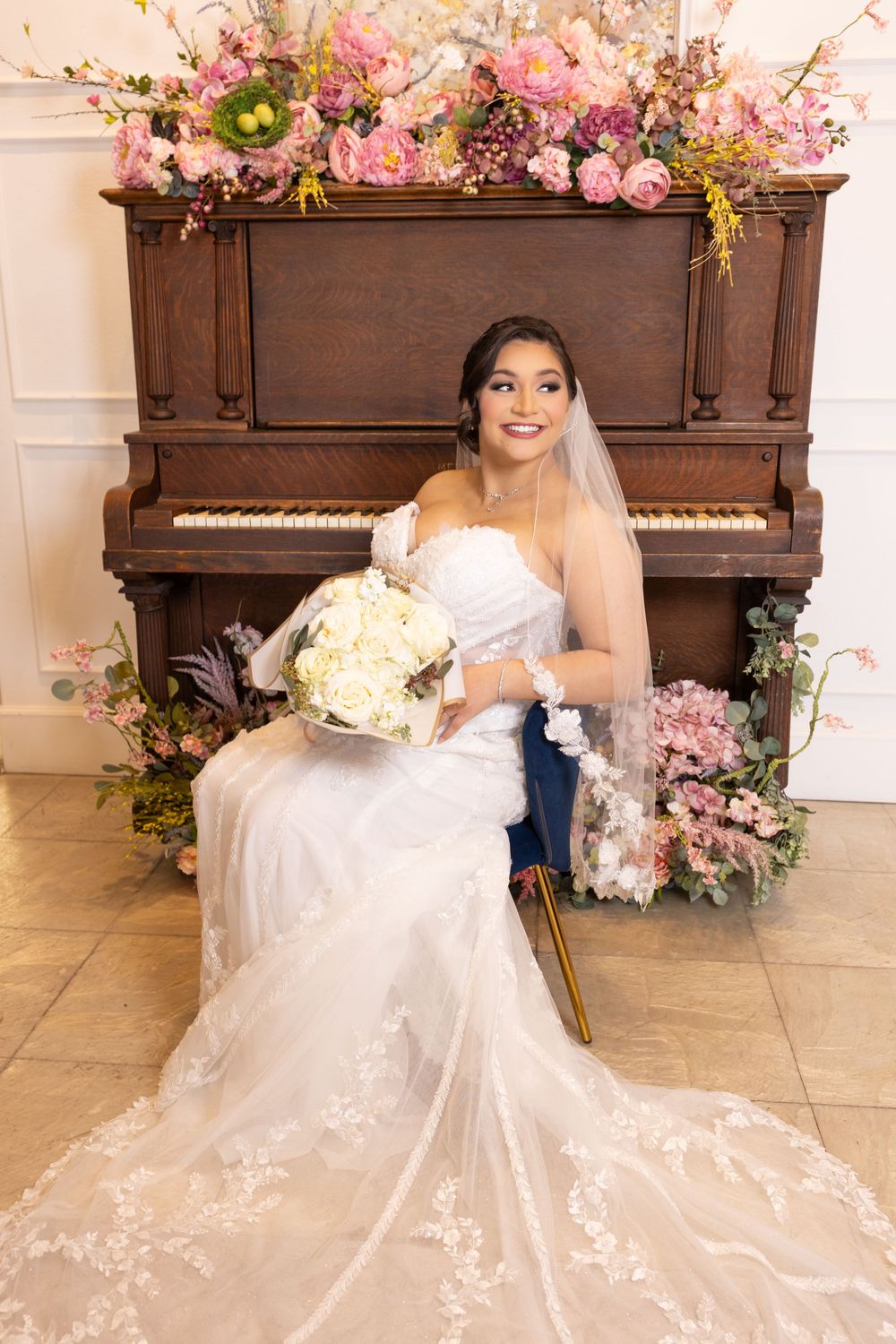 A smiling bride in a white lace wedding dress poses in front of a floral-decorated piano.