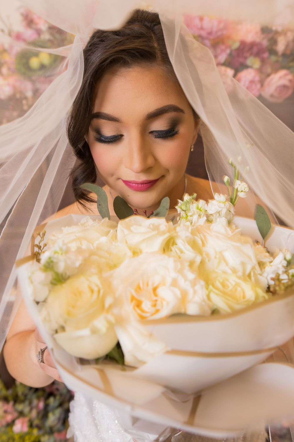 A bride wearing a veil and holding a bouquet of flowers stands against a floral backdrop.