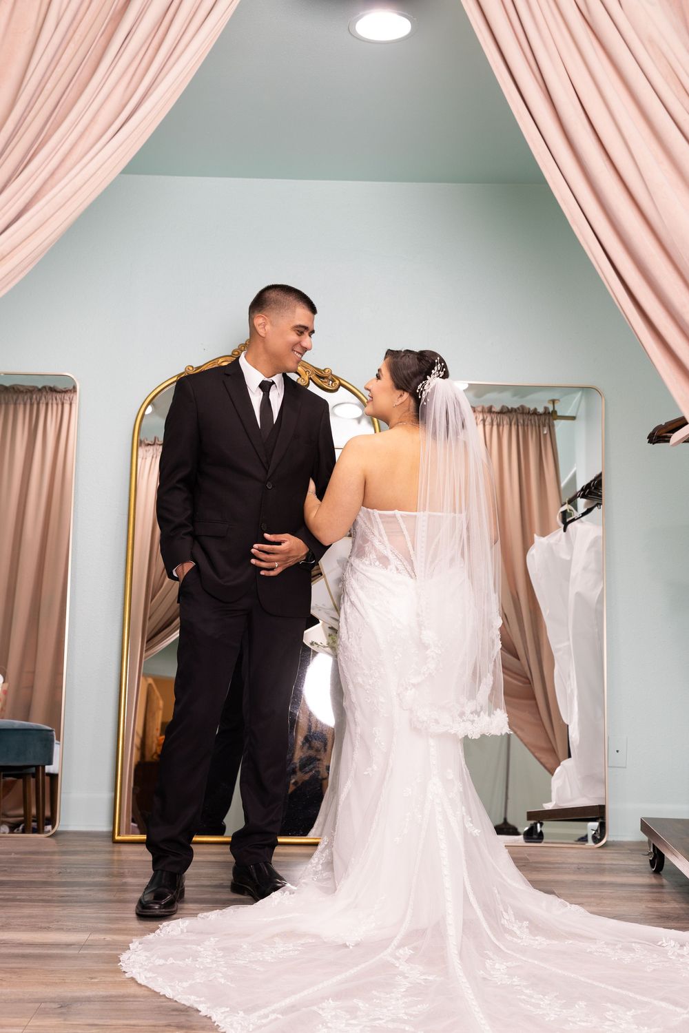 A bride and groom stand together in a room with pink curtains, embracing each other.