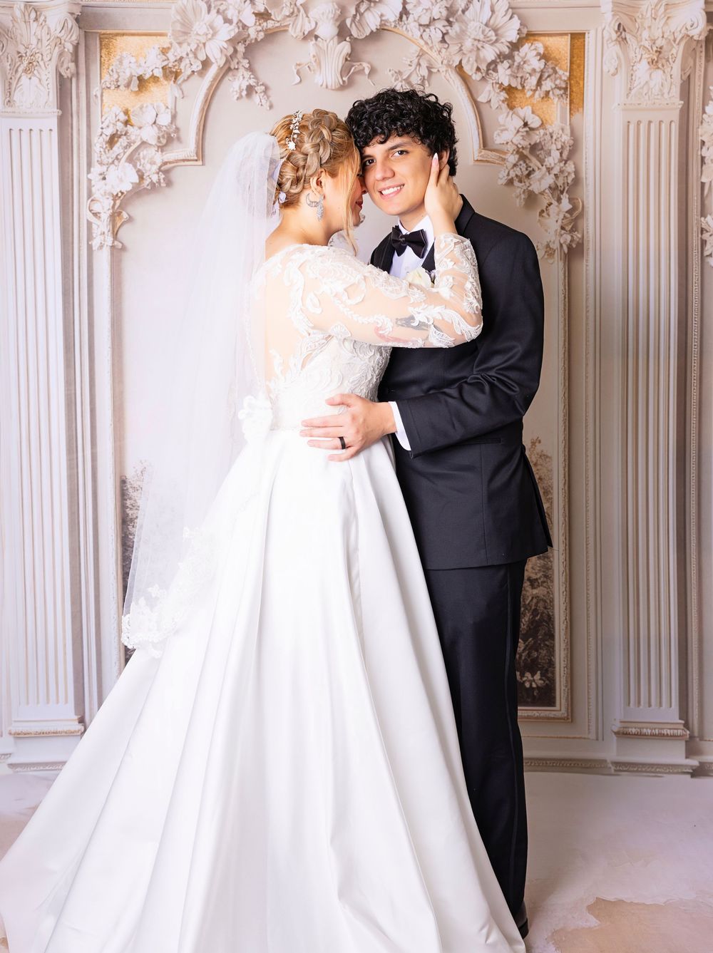 Elegant bride and groom embrace in ornate white venue, bride in lace gown with veil, groom in classic black tuxedo.