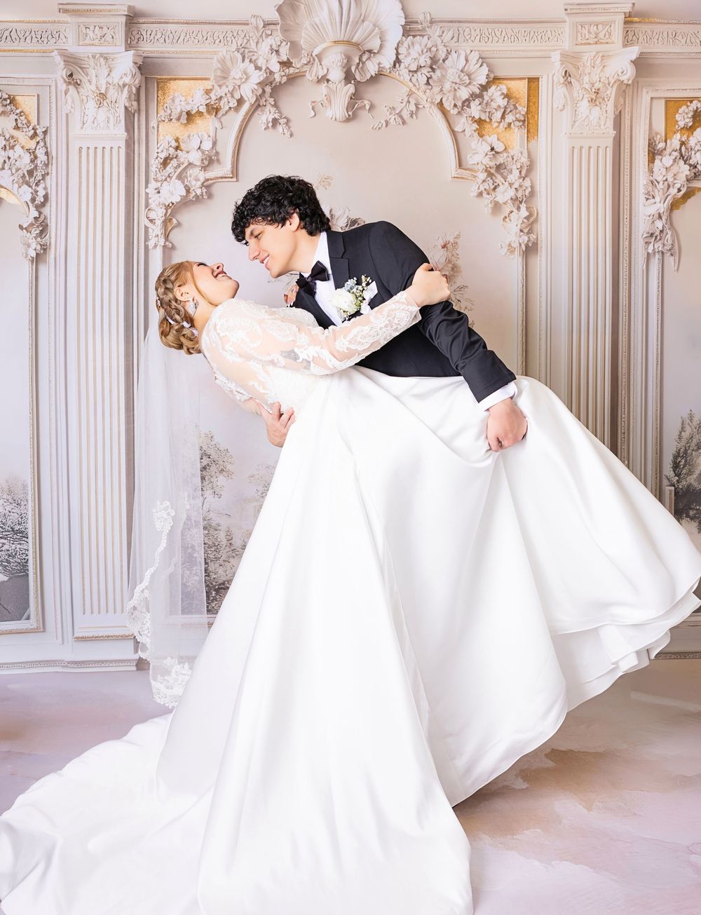 A groom in a black tuxedo dips his bride in a white lace wedding gown in an elegant white ornate venue.