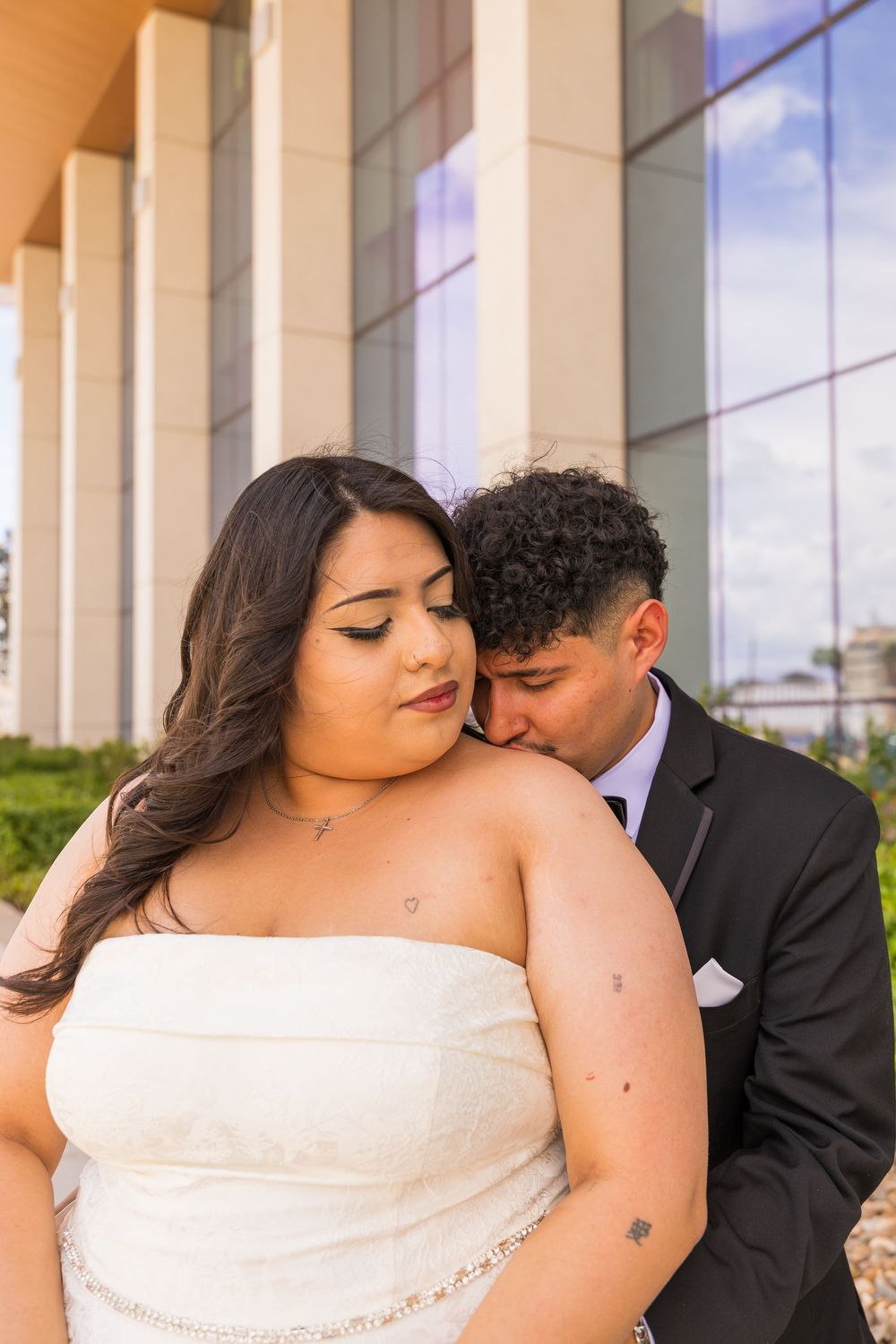A groom in a black suit kisses his bride on the shoulder as she wears a white strapless gown outdoors.