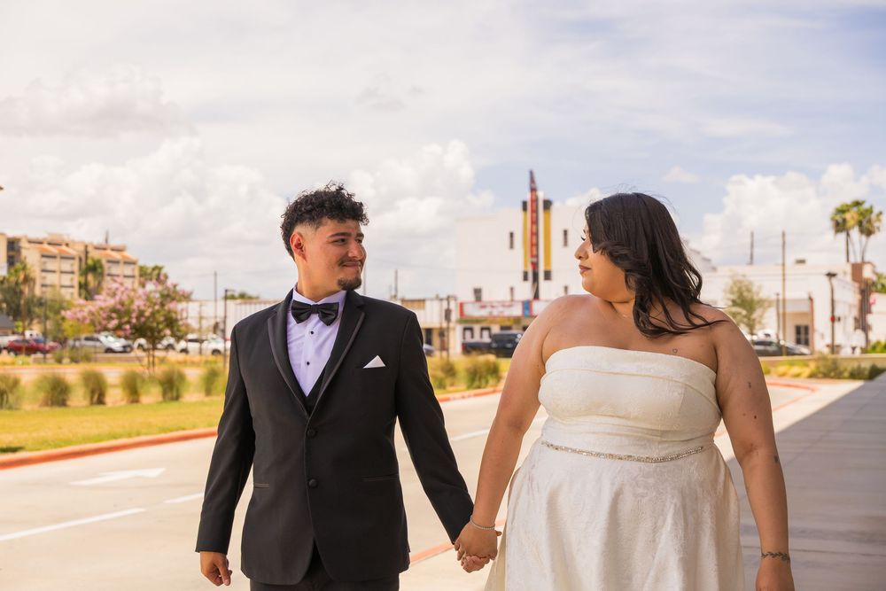 Newlywed couple holding hands in wedding attire, groom in tuxedo and bride in white gown, walking outdoors on sunny day.