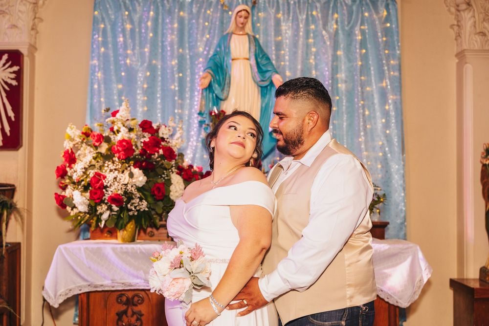 Bride and groom share a romantic moment in a church with a Virgin Mary statue and floral arrangement in the background.