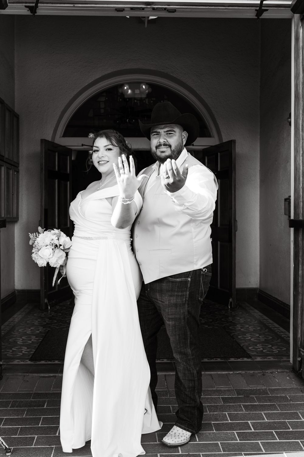 Newlyweds showing wedding rings at church entrance in black and white photo.