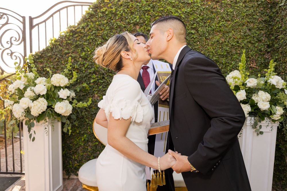 A bride and groom share a romantic kiss under a decorative arch surrounded by white flowers.