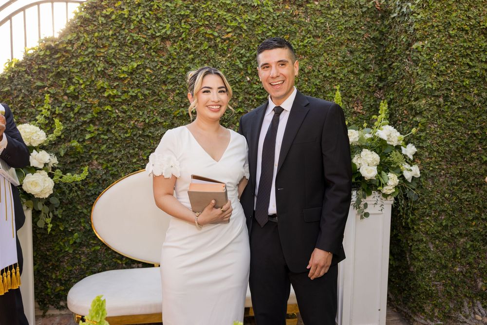 A couple dressed in formal attire stands together in front of a floral arch, celebrating their wedding.