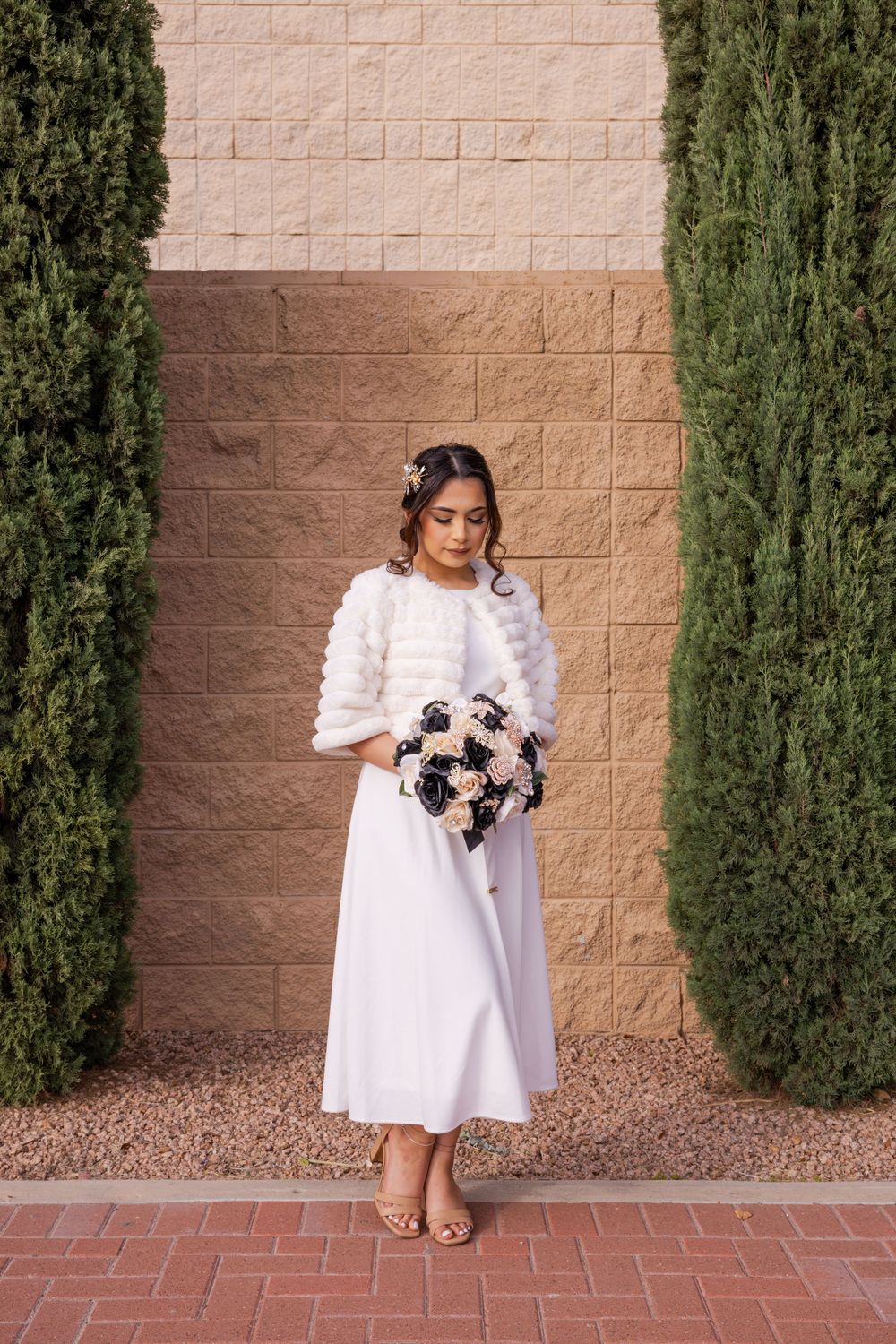 A person in a white midi dress holds a navy and white bouquet while standing between cypress trees on brick pavement.