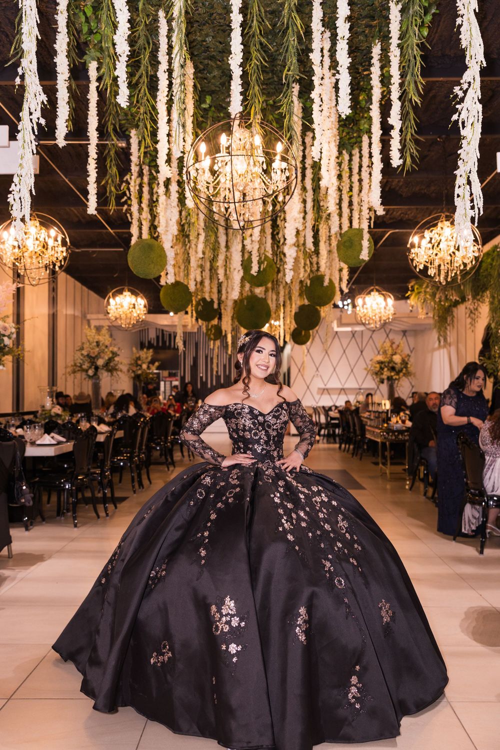 Elegant black ballgowns with floral embellishments displayed in a luxurious venue with hanging chandeliers and greenery.