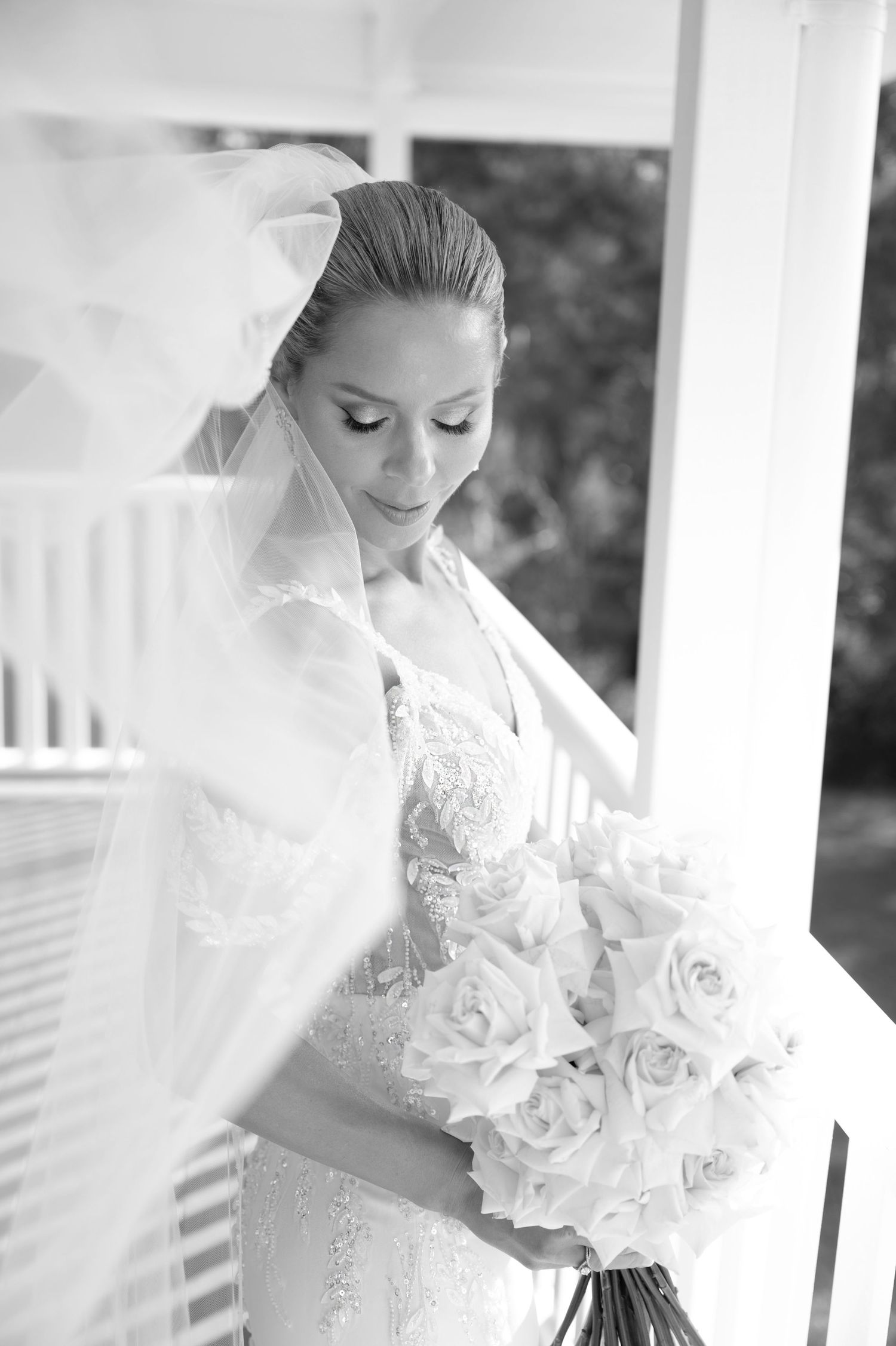 Elegant bride in lace gown holds a white rose bouquet while looking down, captured in black and white photography.