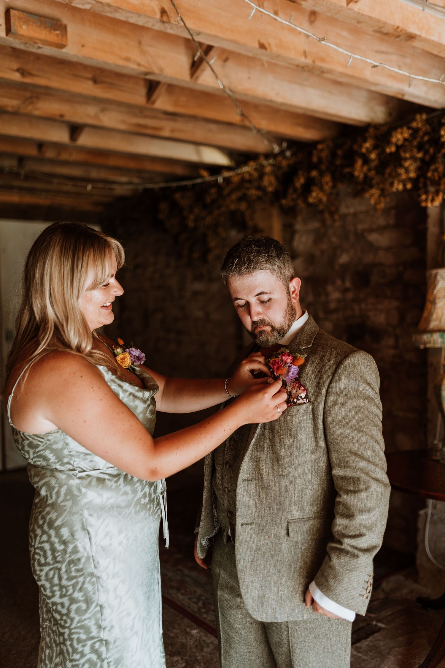 Best woman in a mint green dress pins a boutonniere on groom wearing a gray tweed suit at a rustic indoor venue.