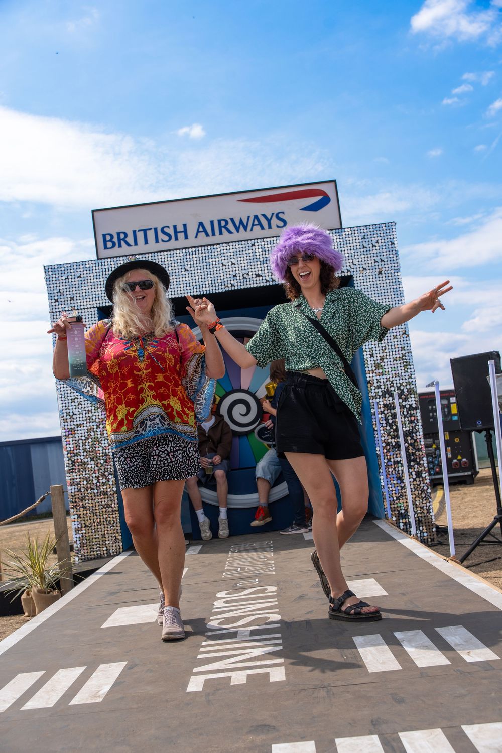 Two people strike fun poses under a British Airways sign at an outdoor festival.