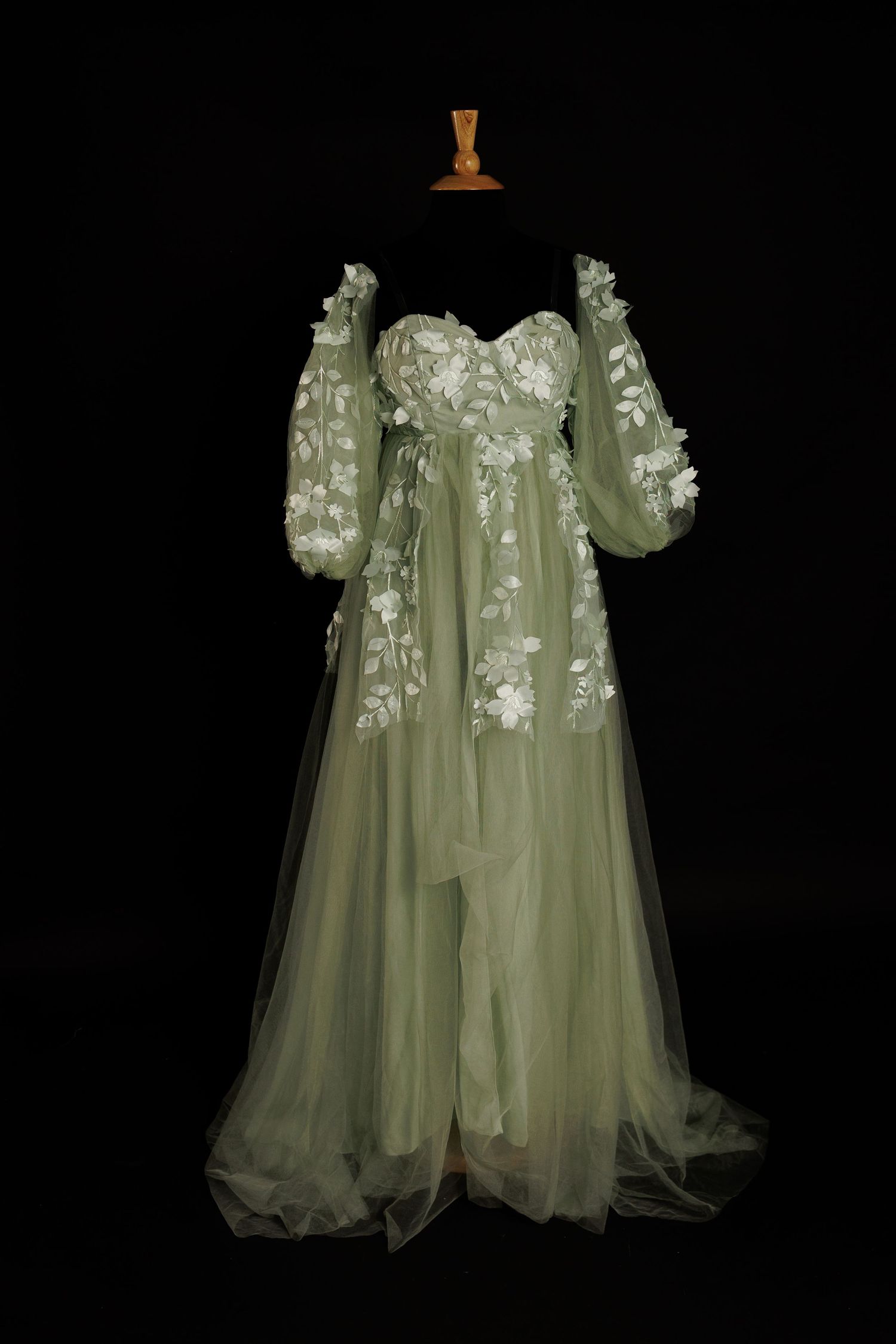 Ethereal sage green tulle dress with delicate white floral embellishments and long flowing sleeves on black background.