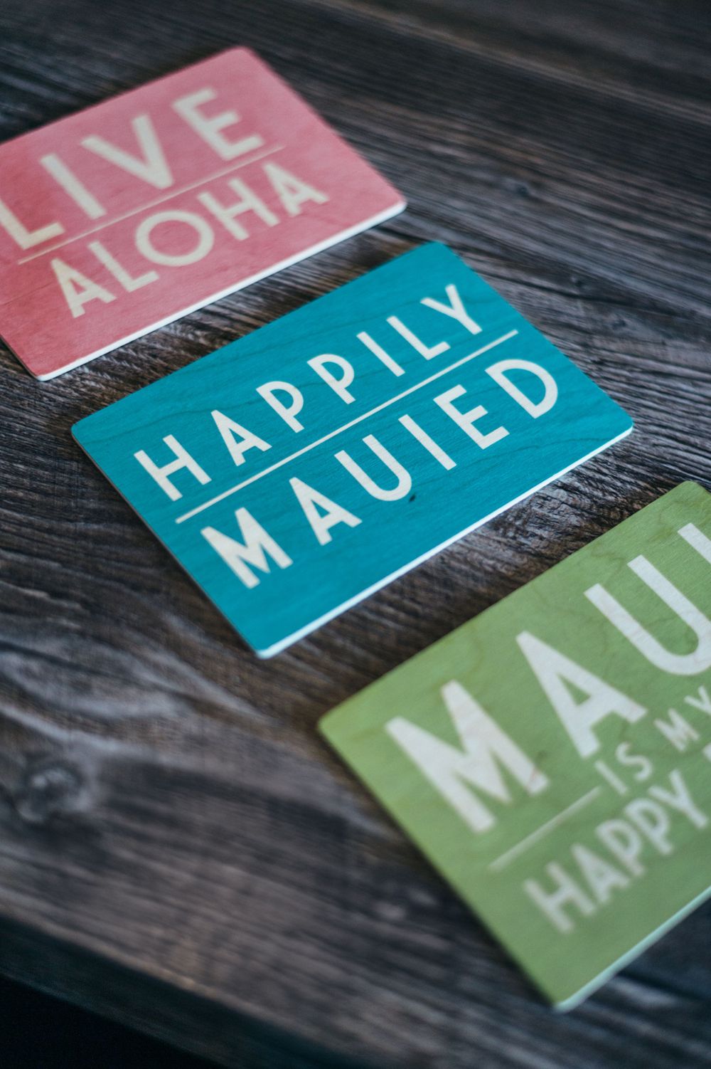 Three colorful Hawaiian-themed signs with phrases including 'Live Aloha' and 'Happily Mauied' on dark wood.
