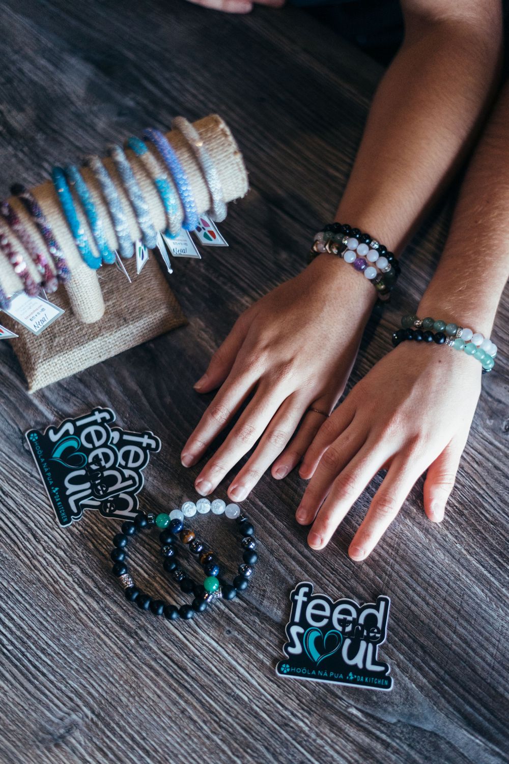 Hands wearing beaded bracelets next to jewelry display and decorative signs on dark wooden surface.