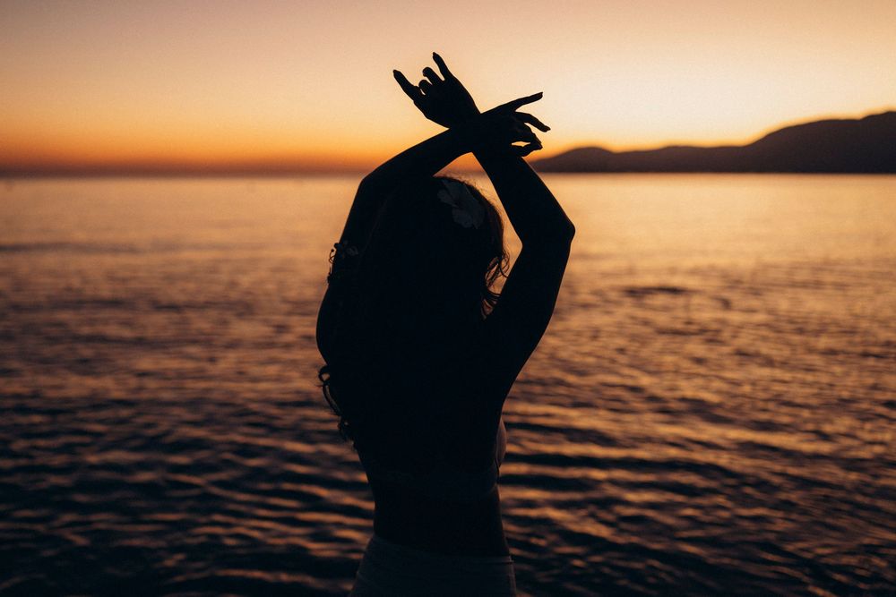 Silhouetted figure raises arms in celebration against vibrant orange and purple sunset over calm ocean waters.