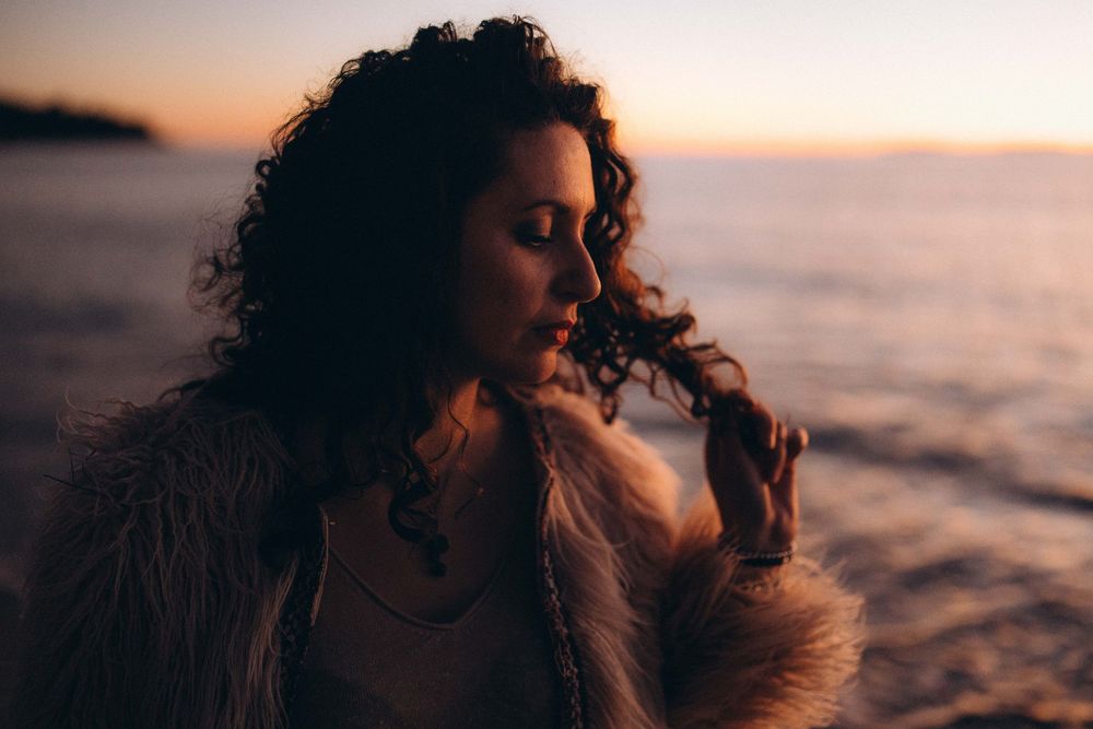 A silhouetted figure with curly hair gazes at sunset by the ocean while wearing a fur-trimmed coat.