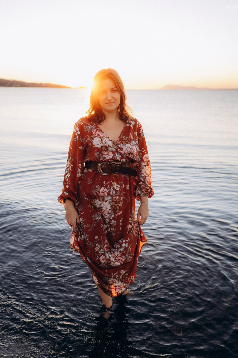 A person in a floral burgundy top poses by the ocean at sunset with glowing golden light.