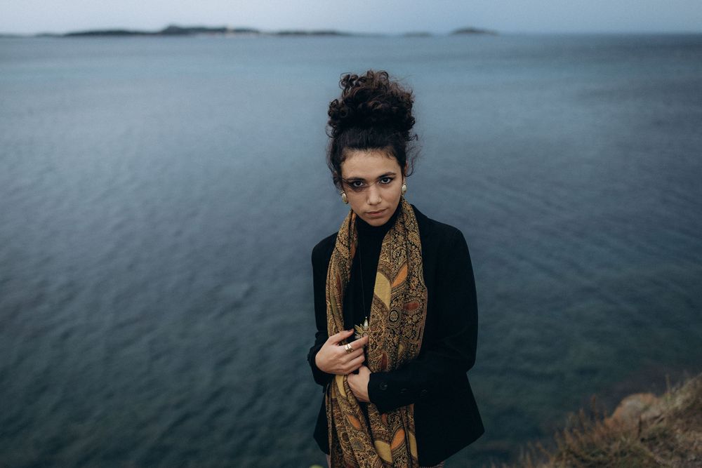 A person in a black coat and brown scarf stands on a rocky cliff overlooking a moody gray ocean.