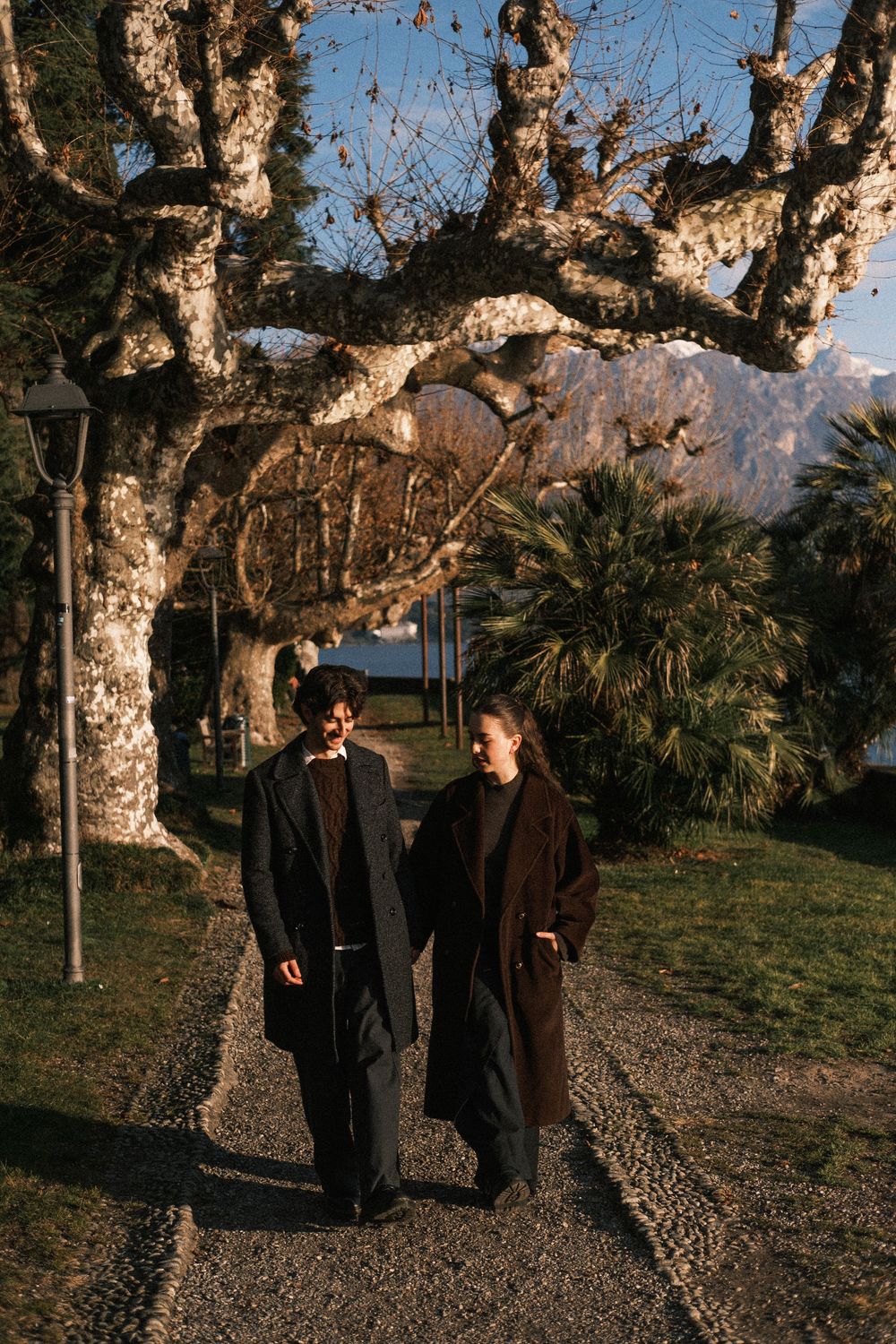 People in dark coats walk along a gravel path beneath bare trees during golden hour with mountains in the background.