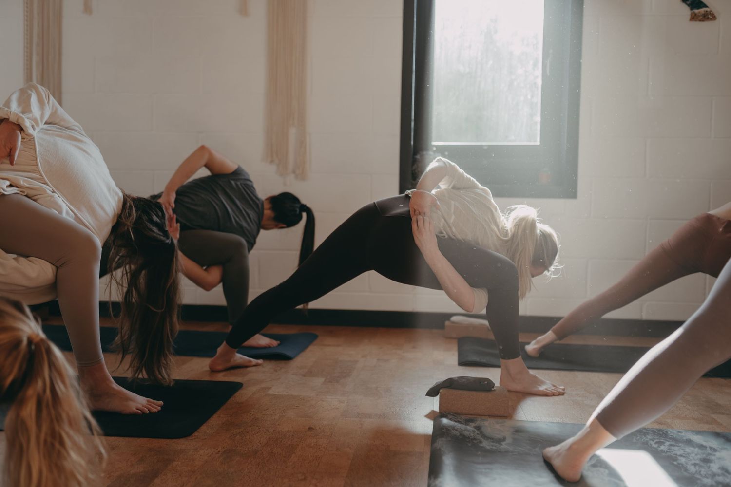 A yoga class practices poses on white mats in a bright studio with natural light streaming through the windows.