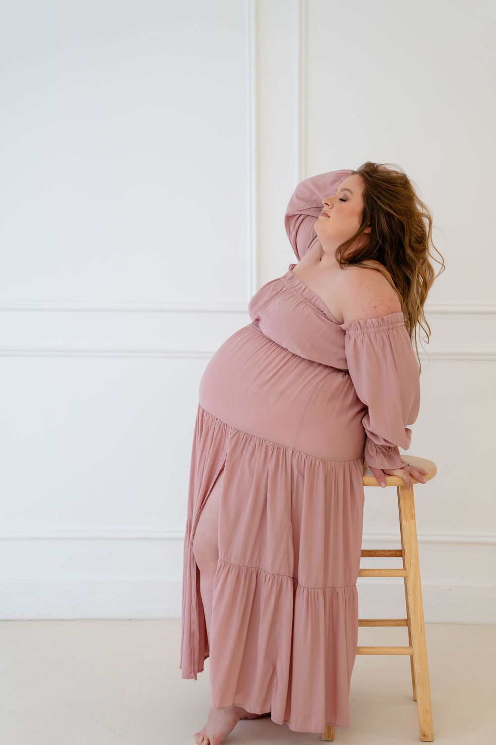 Artistic maternity portraits in a flowing dusty rose dress against white walls.